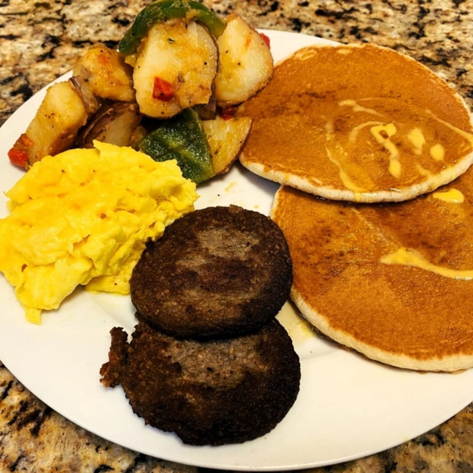 A Gutbusters Breakfast plate with scrambled eggs, home fries, sausage patties, and pancakes with syrup.