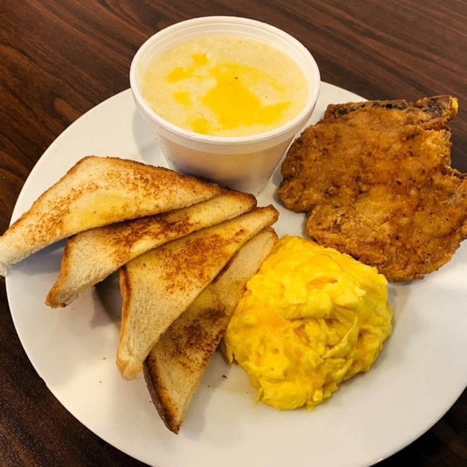 A Gutbusters Breakfast plate with fried pork chop, scrambled eggs, buttered toast, and grits.