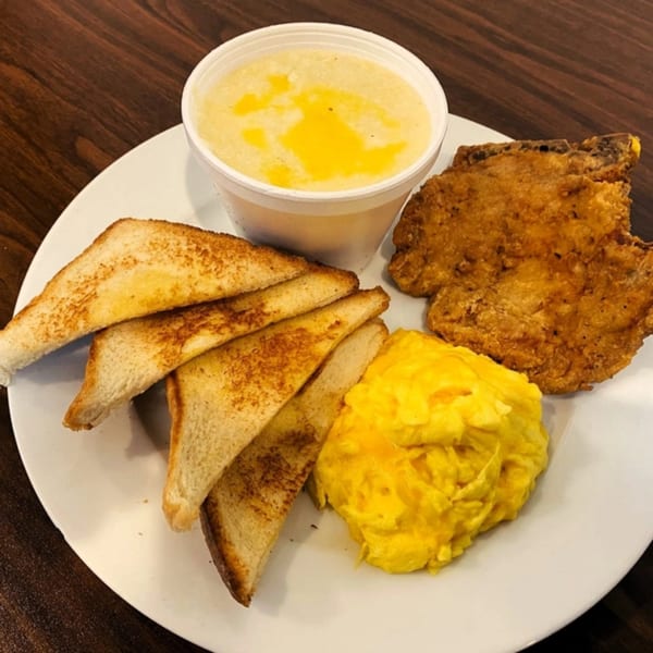 A Gutbusters Breakfast plate with fried pork chop, scrambled eggs, toast, and a cup of grits.