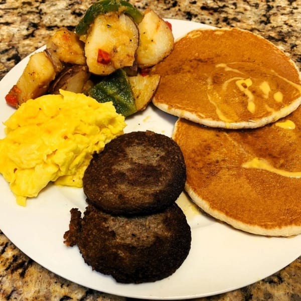 A Gutbusters Breakfast plate with scrambled eggs, home fries, sausage patties, and pancakes with syrup.