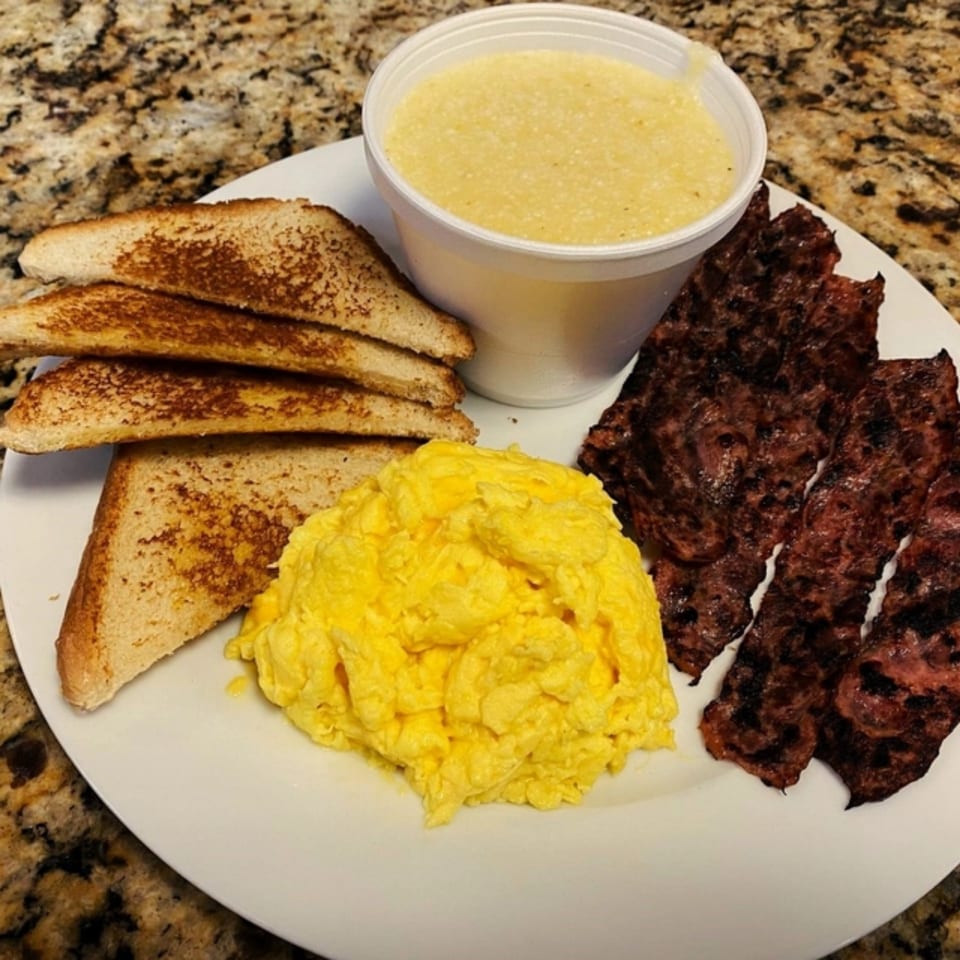 A Gutbusters Breakfast plate with scrambled eggs, toast, crispy bacon, and a cup of grits.